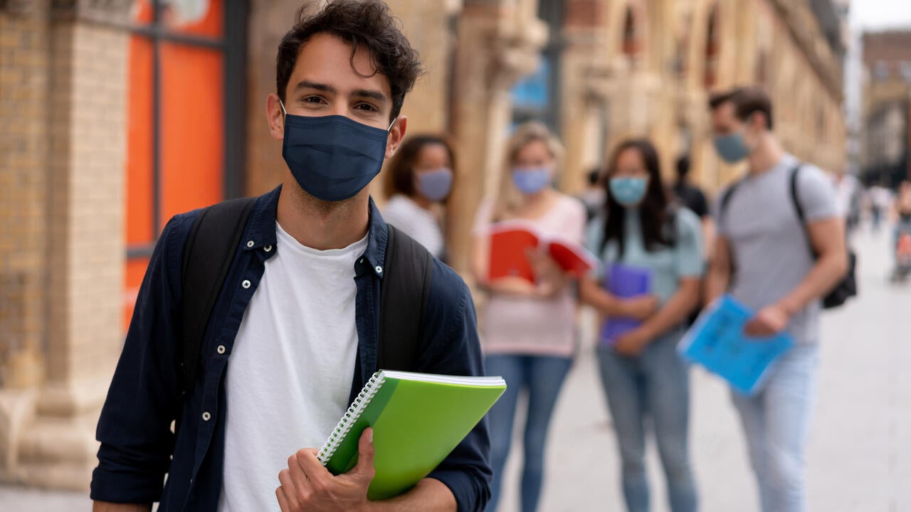 A student wearing a face mask and holding a book with other students in the background