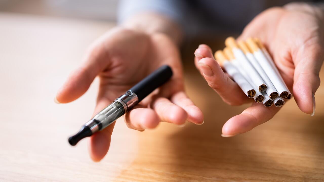 Person holds an e-cigarette in one hand and a pile of cigarettes in the other.