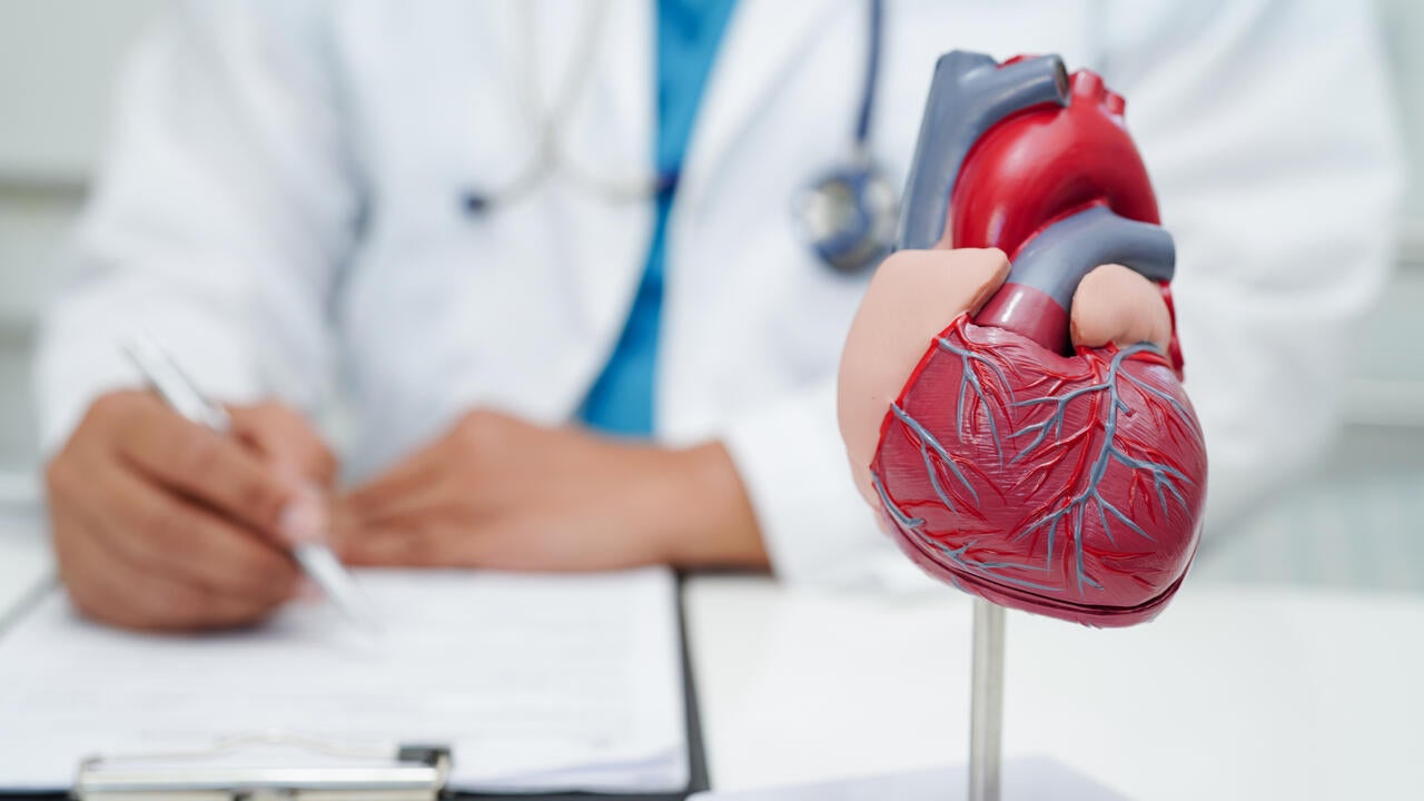 Doctor making notes on clipboard on desk with model of a human heart in foreground.