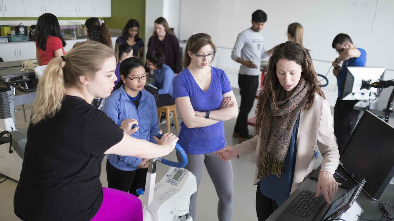 Students gathered around an instructor pointing to data on a screen, with one student on a stationary bike