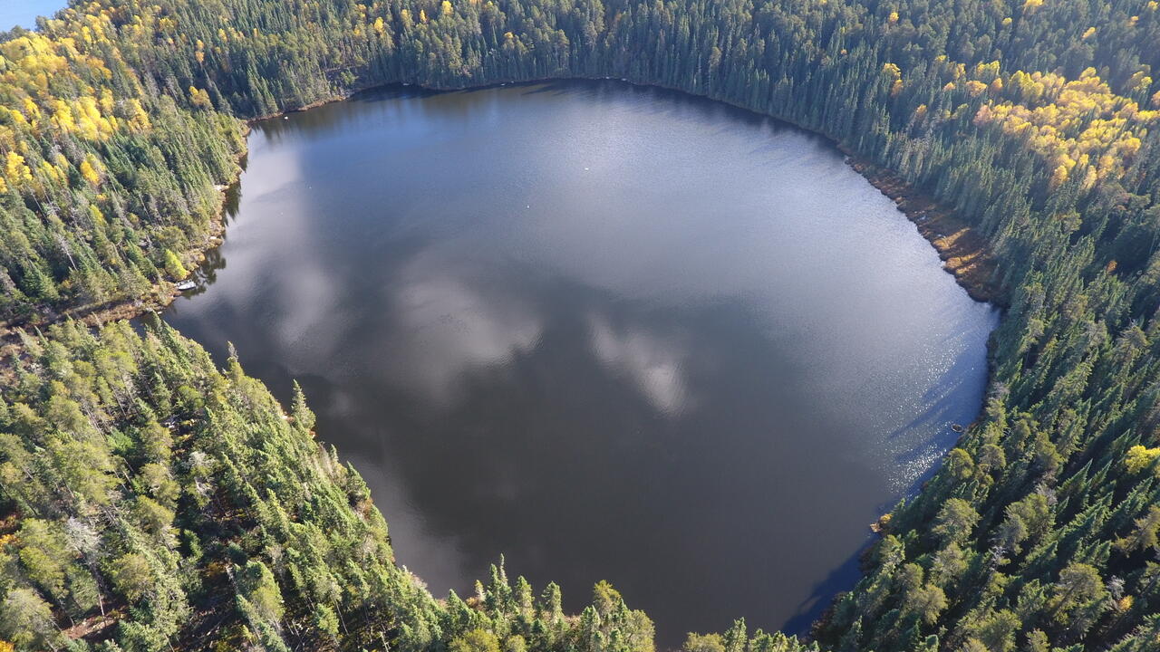 A birds eye view of a dark lake surrounded by green spruce trees
