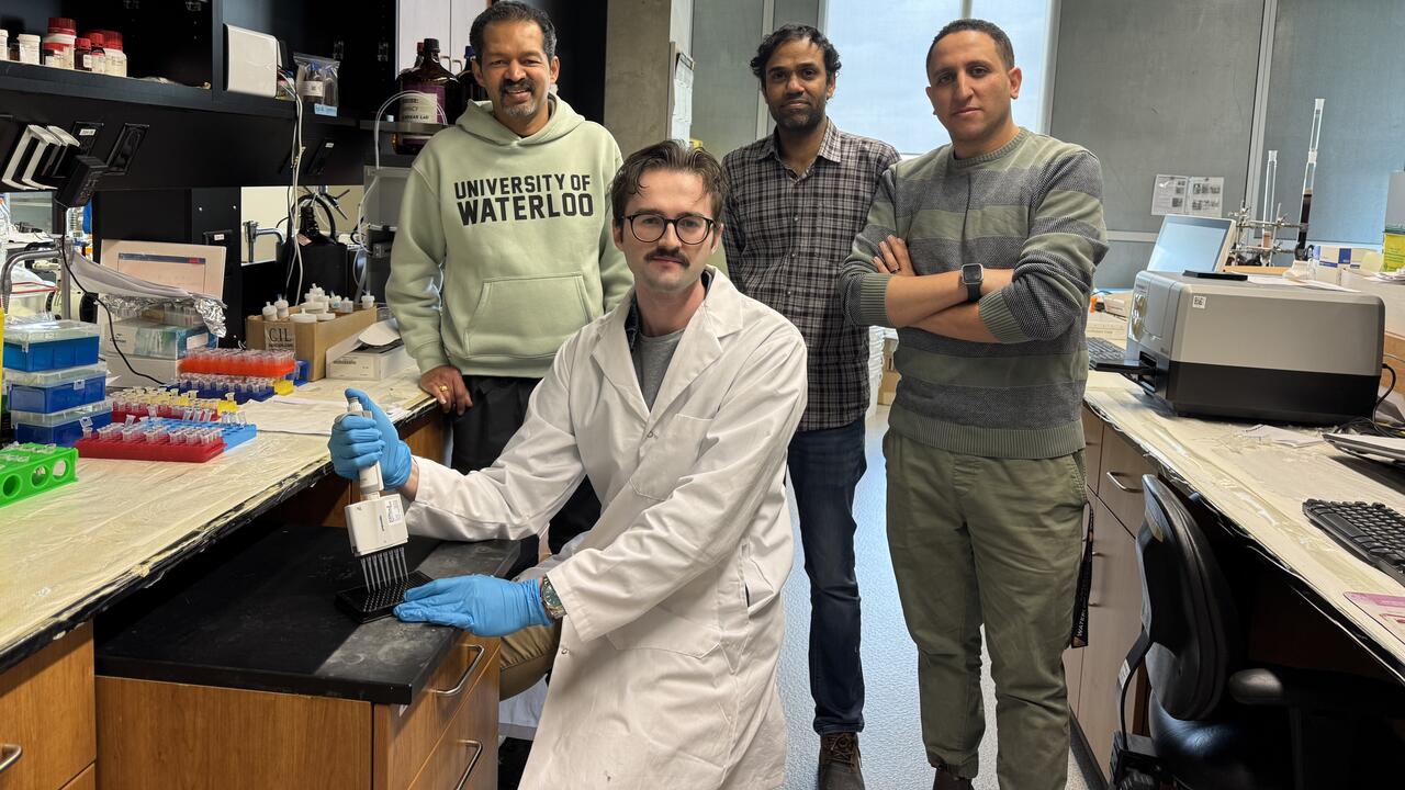 Four people in a lab, with one of them seated and wearing a lab coat and gloves