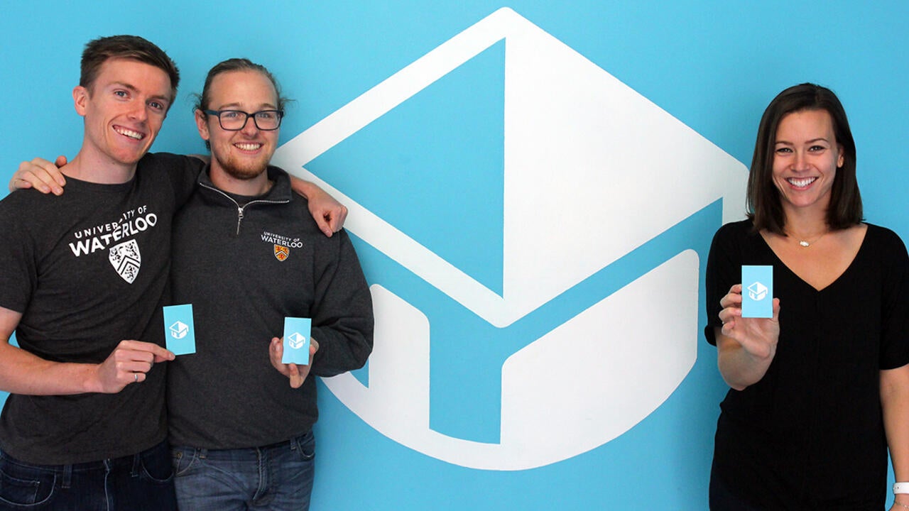 Jack Forbes, Zach Lima, and Courtney Sabo standing with business cards in front o company logo