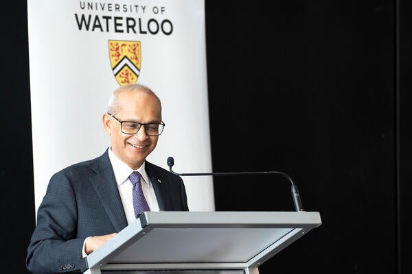 President Goel smiles as he reviews notes on a podium in front of a UWaterloo banner.