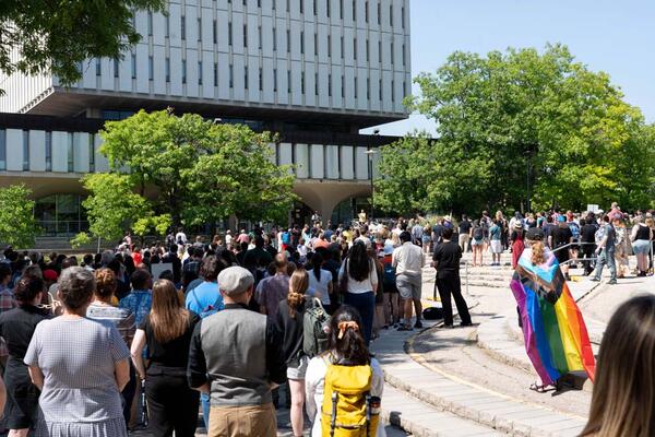 A crowd is assembled in the Arts Quad listening to speakers presenting from steps of Dana Porter Library. 