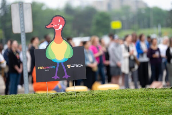 Pride signage featuring a rainbow goose is pictured on a lawn in front of a large group assembled in lot H