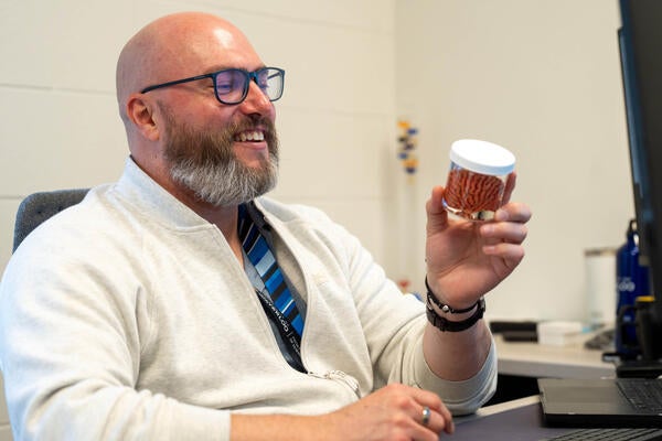 Travis Craddock examines a miniature model of a brain in a jar while seated at his desk.