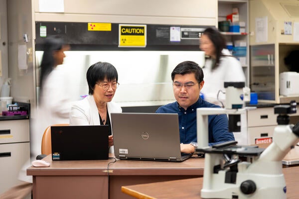 Helen Cho and Bing Ho work on computer in lab. People are blurred as they walk behind them.