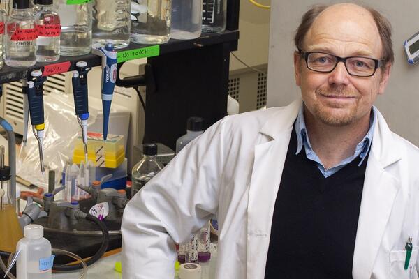 Professor wearing lab coat stands in front of a shelf of beakers in a laboratory