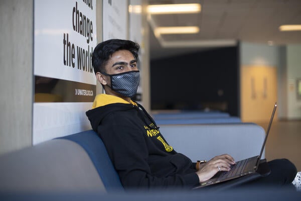 A student sitting in a Waterloo hoodie and mask while typing on a laptop