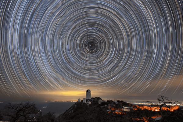 Circles of light on the night sky. A telescope dome atop a mountain is below the center of the circle.