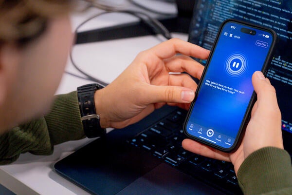 Rastin Rassoli holding a mobile phone with screen showing Doro AI interface and a laptop with coding in the background