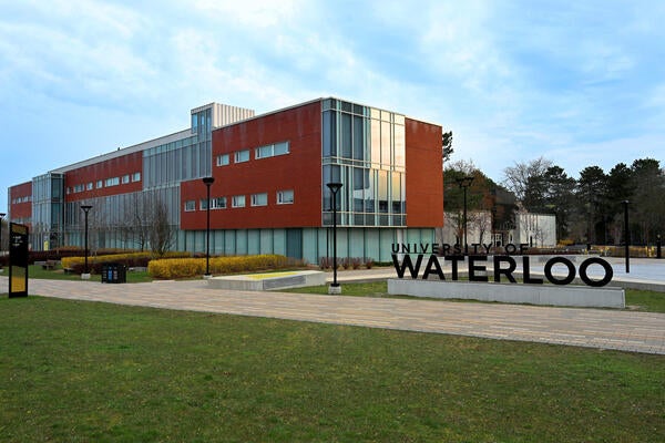 University of Waterloo signage in the foreground with Tatham Centre in the background on a summer cloud filled day