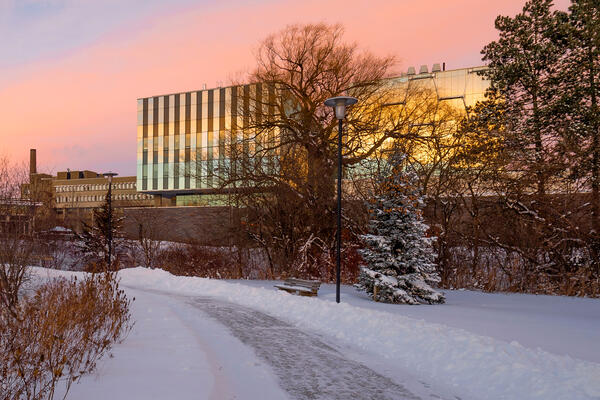 A snowy pathway leads through a winter landscape, with the QNC building reflecting the colors of a sunset in the background.