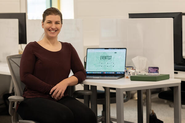 Ciara Azam sitting by a desk with a laptop showing the Eventist landing page next to a box of tissues and trinkets