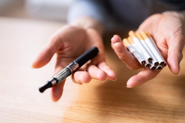 Person holds an e-cigarette in one hand and a pile of cigarettes in the other.