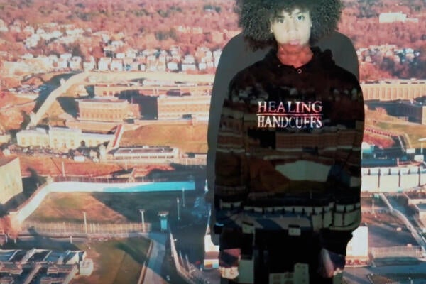 A Black teenager wearing a hoodie that says "Healing Handcuffs" stars at the camera, an image of a prison projected across him