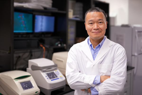 Juewen Liu is wearing a white lab coat and blue collared shirt. He is standing with his arms crossed and smiling in his lab.