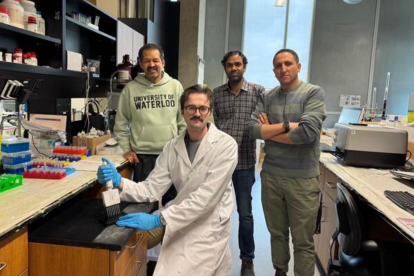 Four people in a lab, with one of them seated and wearing a lab coat and gloves
