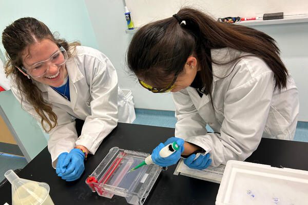 Science students conducting an experiment in a classroom. They are wearing white lab coats, goggles, and blue gloves.