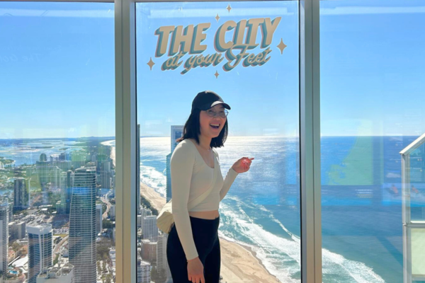 Tiffany Chan standing in a tall building with glass windows pointing to the beach shoreline in Australia. 