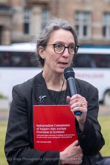 Alison Phipps is dressed in all black clothing, holding microphone in one hand and a red book in the other