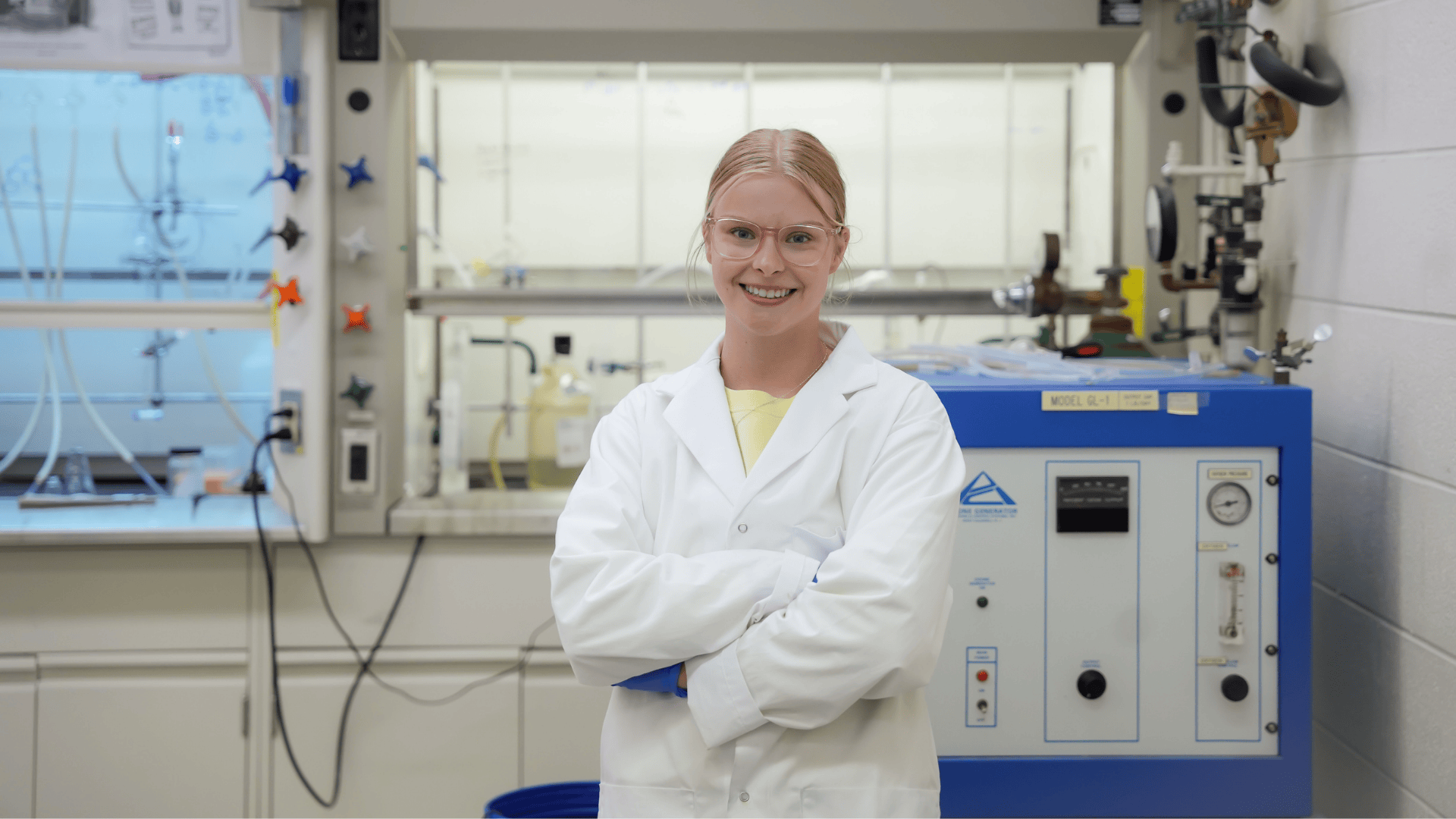 Allysa wearing safety glasses and a white lab coat stands confidently with arms crossed in a chemistry lab.