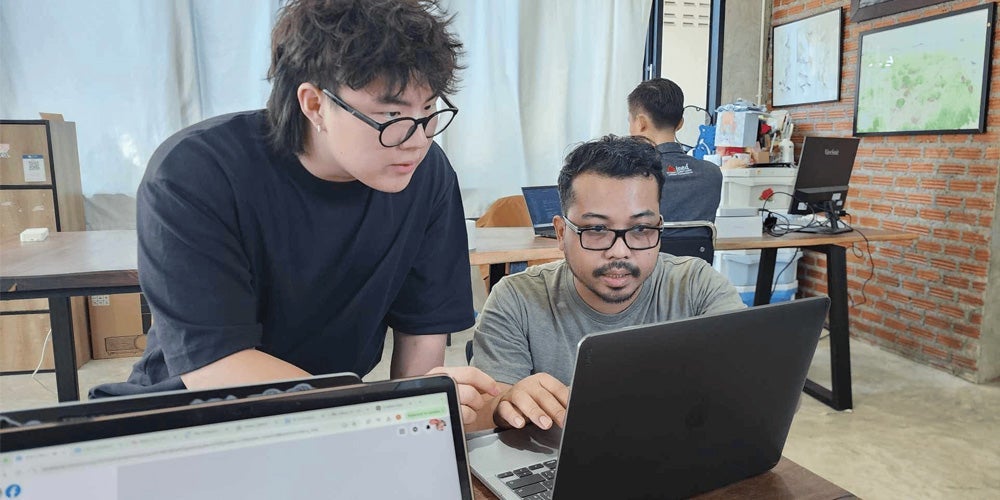 Two young men work together on a laptop at a wooden table in an office setting, with another person working at a computer in