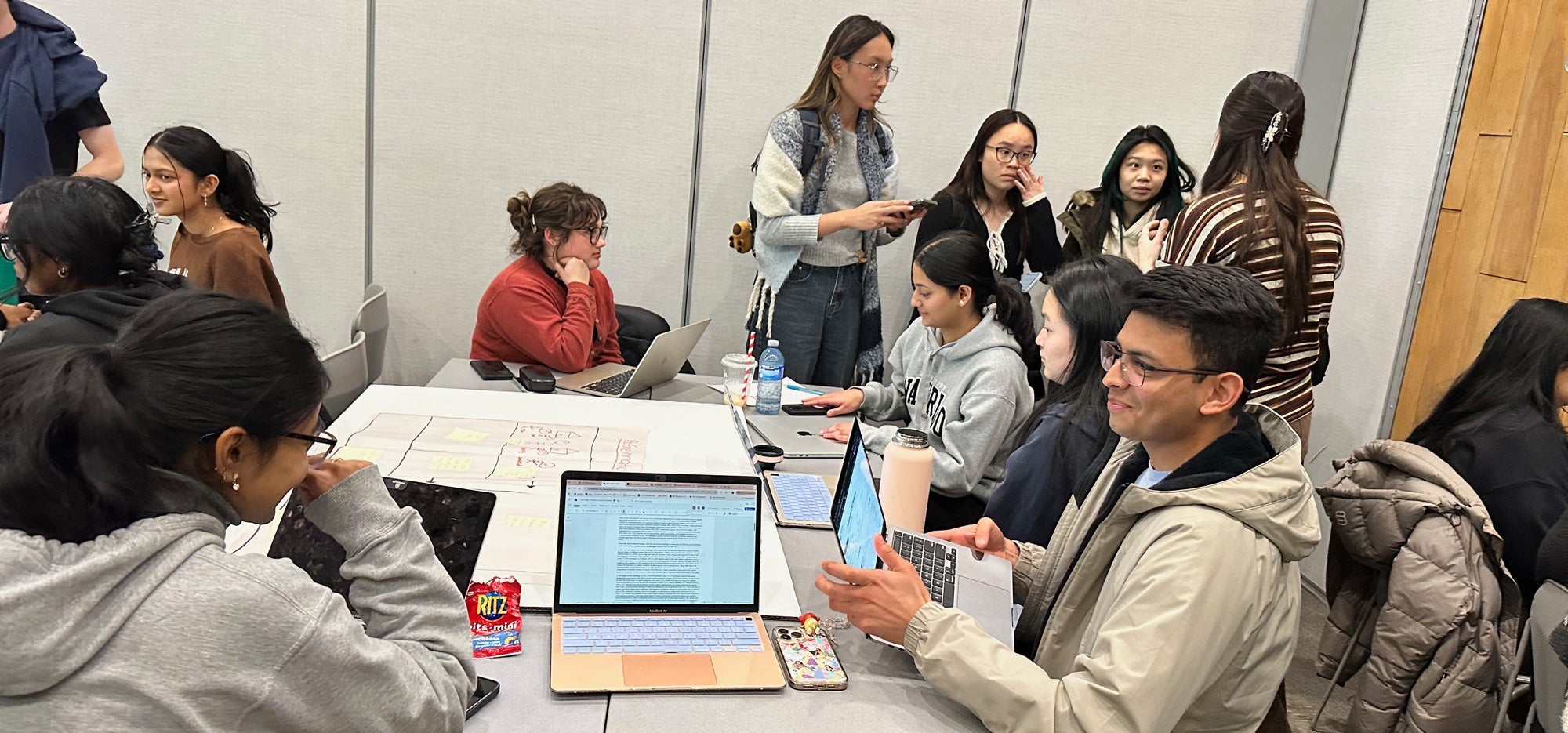 Students around a table with laptops and others in the background