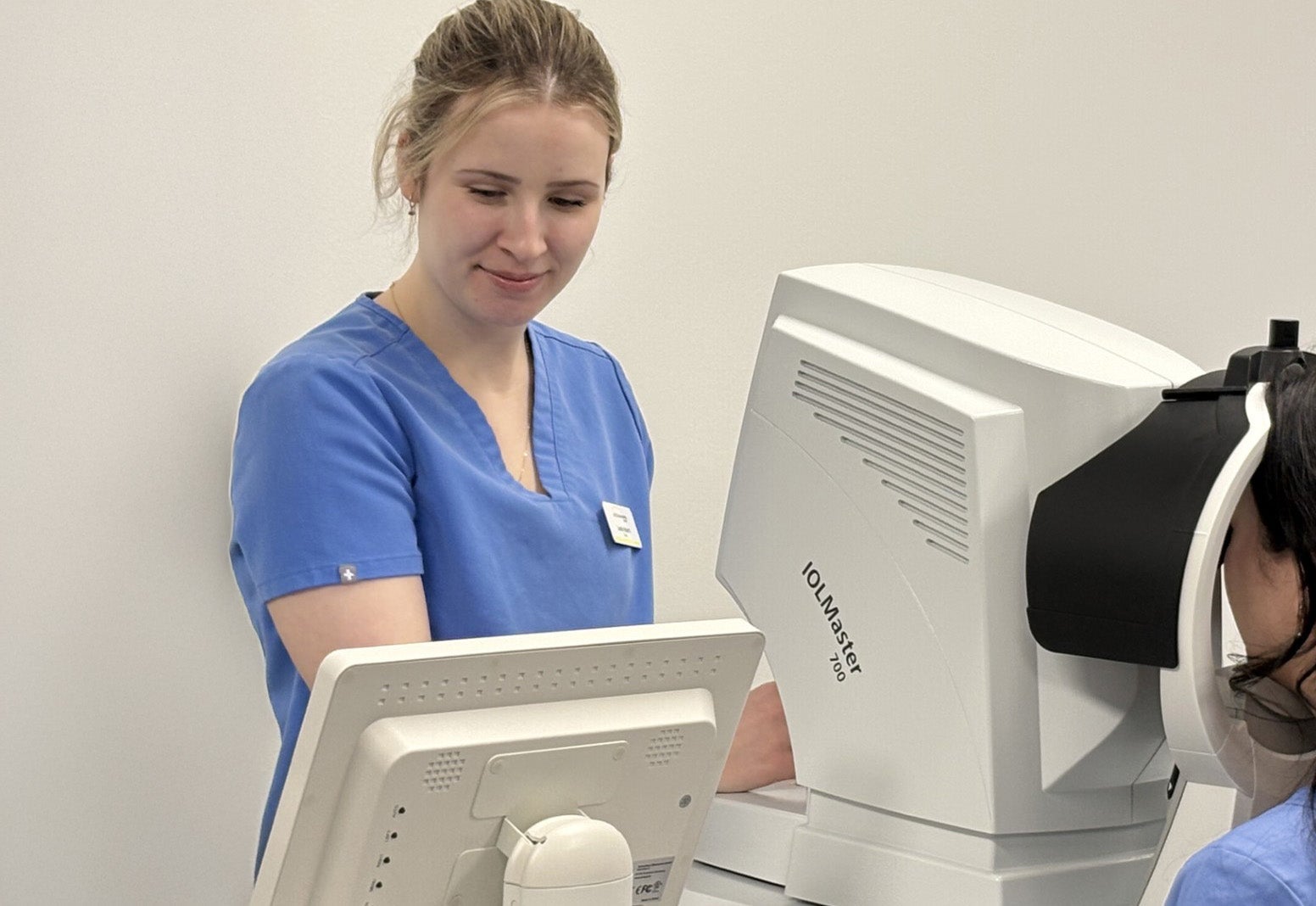 A healthcare professional in blue scrubs is operating an IOLMaster 500 device to conduct an eye examination on a patient.