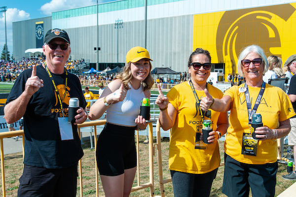 Four people dressed in black and gold holding drinks in one hand and giving thumbs up or number one with the other