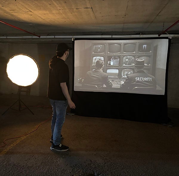 The artist stands infront of a screen projecting an archival image of two security guards watching a wall of 16 monitors.