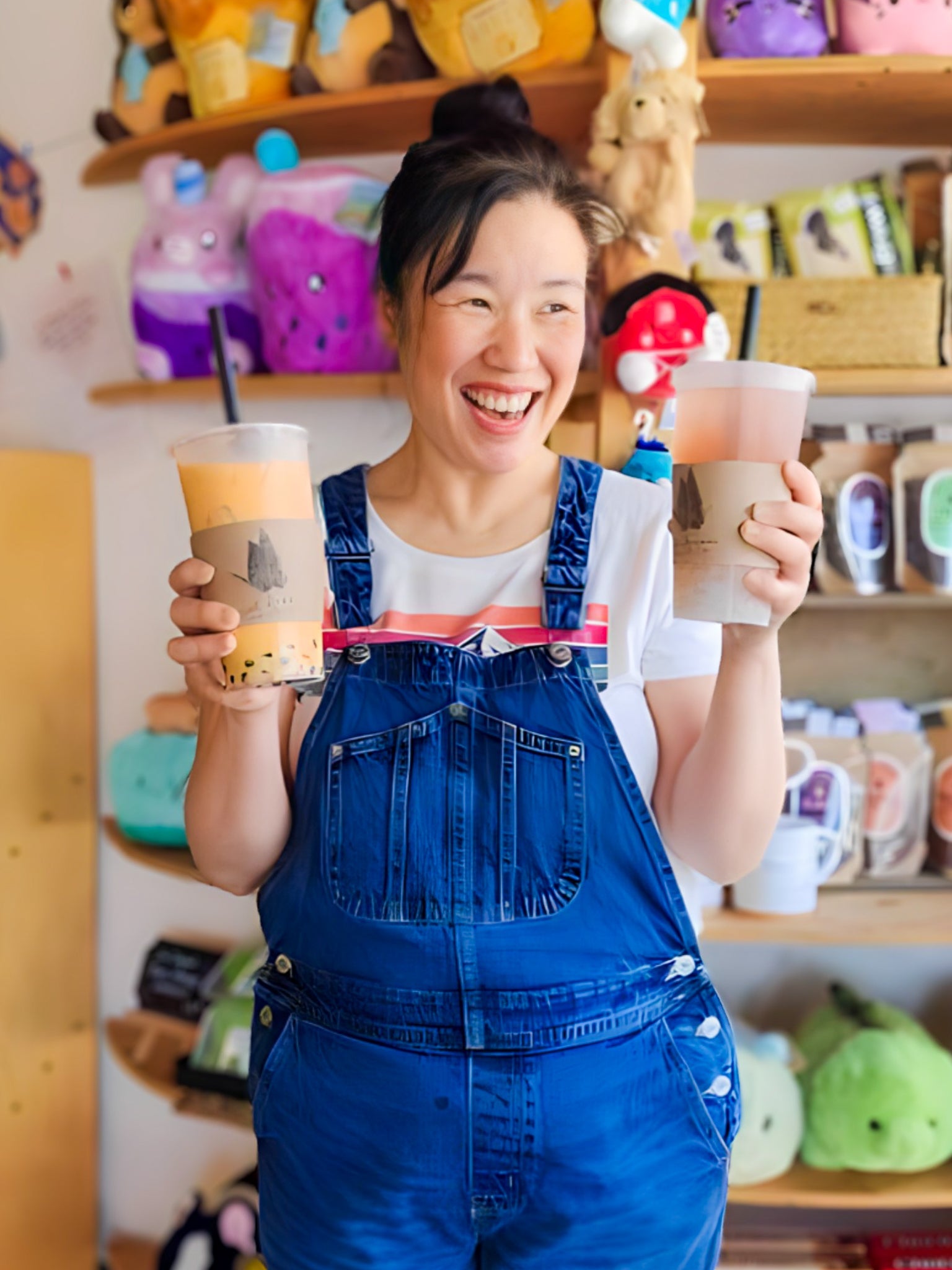 Jin-Hee (Jinny) Song holding bubble tea in each hand and smiling in front of merchandise wall inside Sweet Dreams Teashop