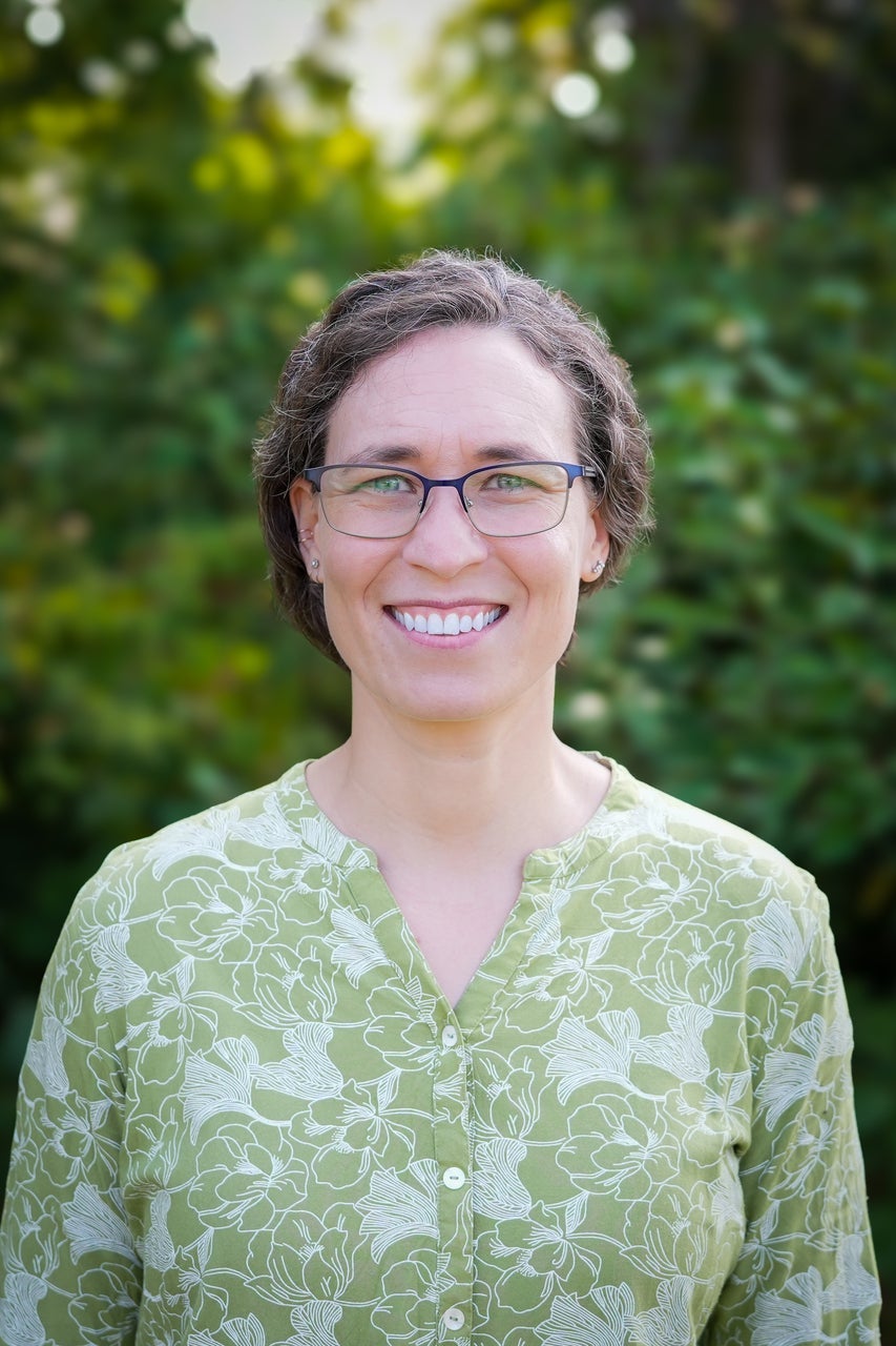 Julie Messier is wearing a green and white floral shirt, is smiling and is against a backdrop of outdoor greenery.
