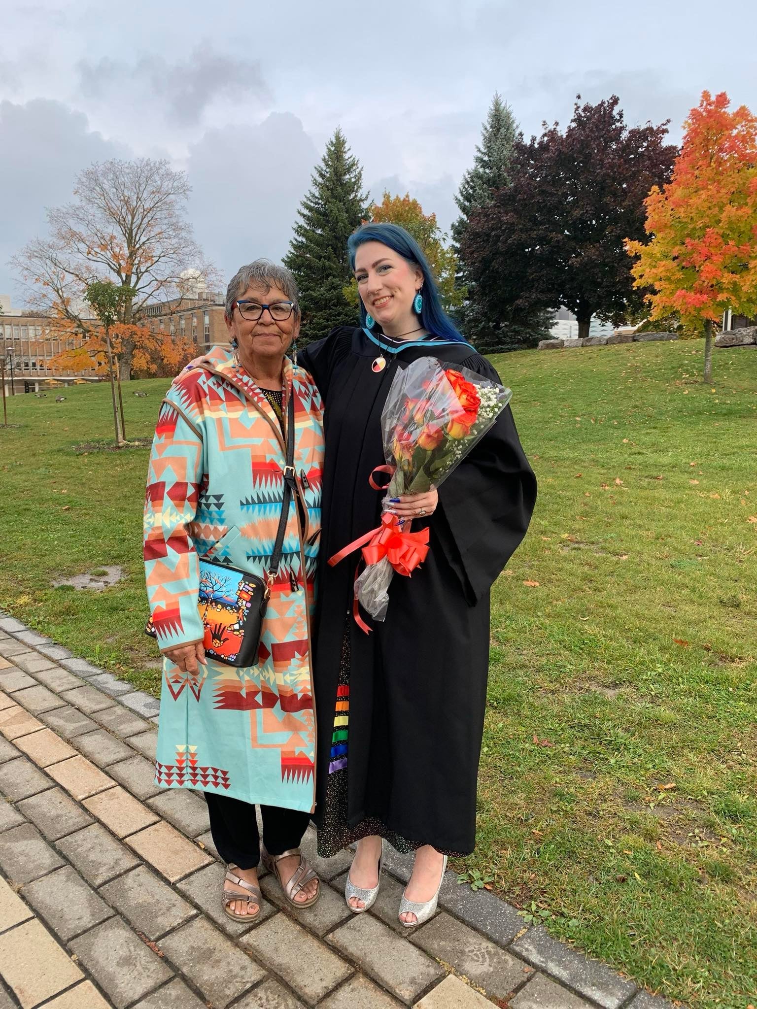 Kaylee wearing a graduation gown and holding flowers. She is standing next to her kôhkom.
