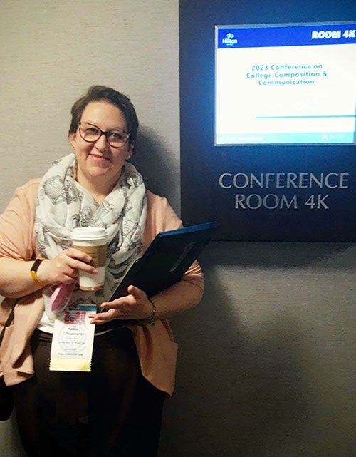 Kellie Chouinard with a coffee cut and a lap top, next to a sign pointing to a conference room