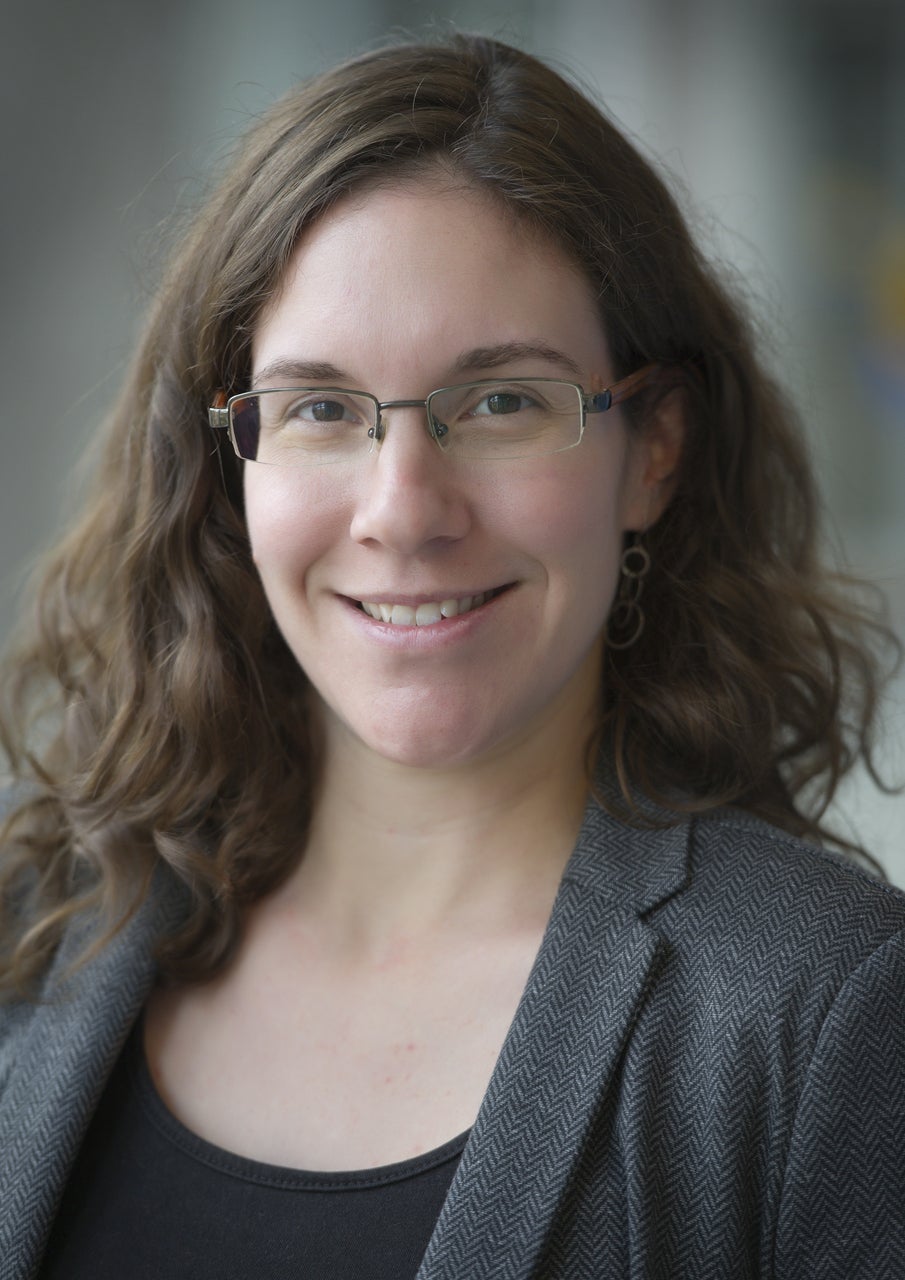 Academic headshot of Laura Hug, smiling, with long wavy brown hair, glasses, and a grey blazer over a dark top.
