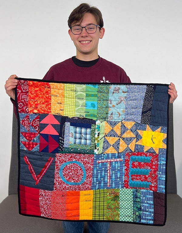 Lucas holds up a small quilted wallhanging with rainbow colours, embroidery and quilted motifs described in the article