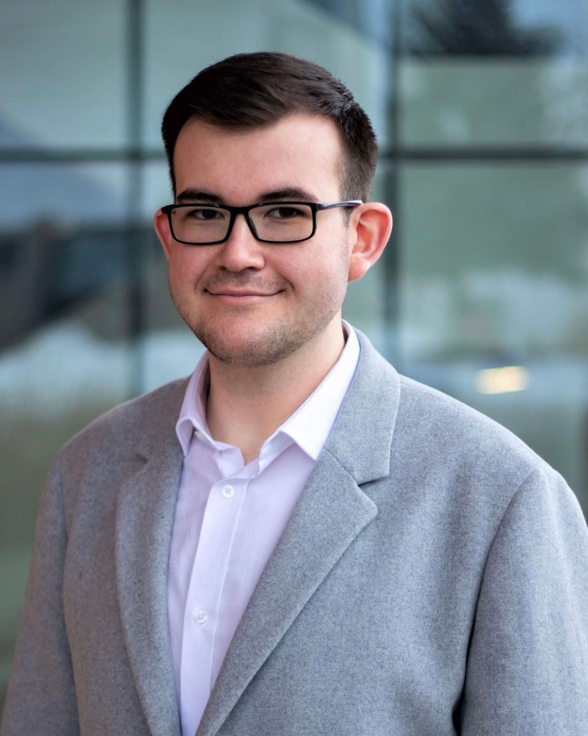 Nikola Cubelic is wearing a white collared shirt, a grey blazer and black rimmed glasses.