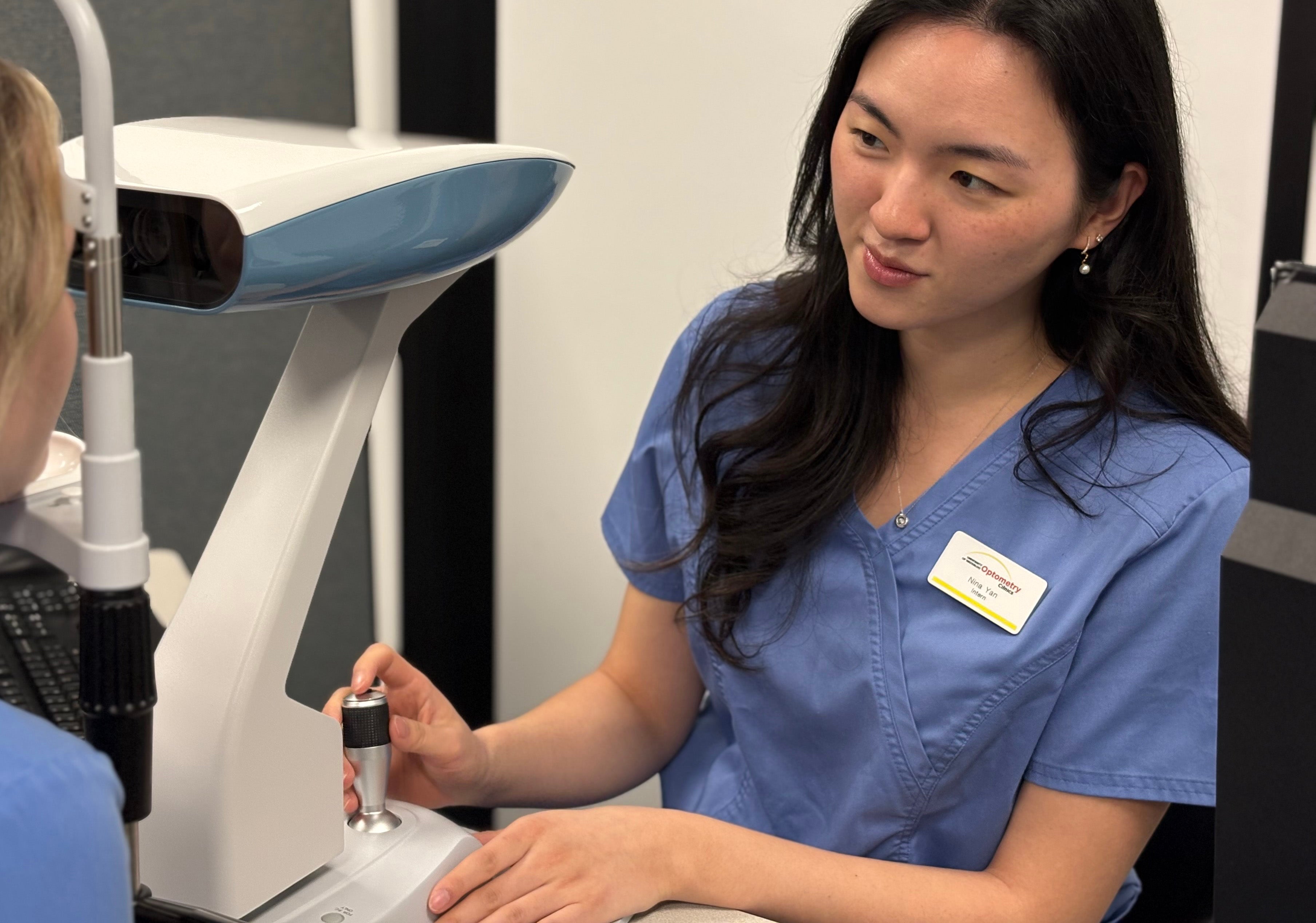 A healthcare professional in blue scrubs is operating an ophthalmic device to perform an eye examination on a patient.