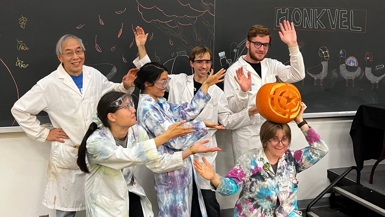 Six scientists wearing white and tie dye lab coats posing silly in front of a blackboard.