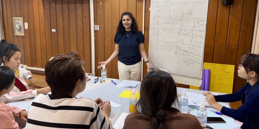A young  stands in front of a whiteboard covered with mathematical equations, explaining to a group of seated people.