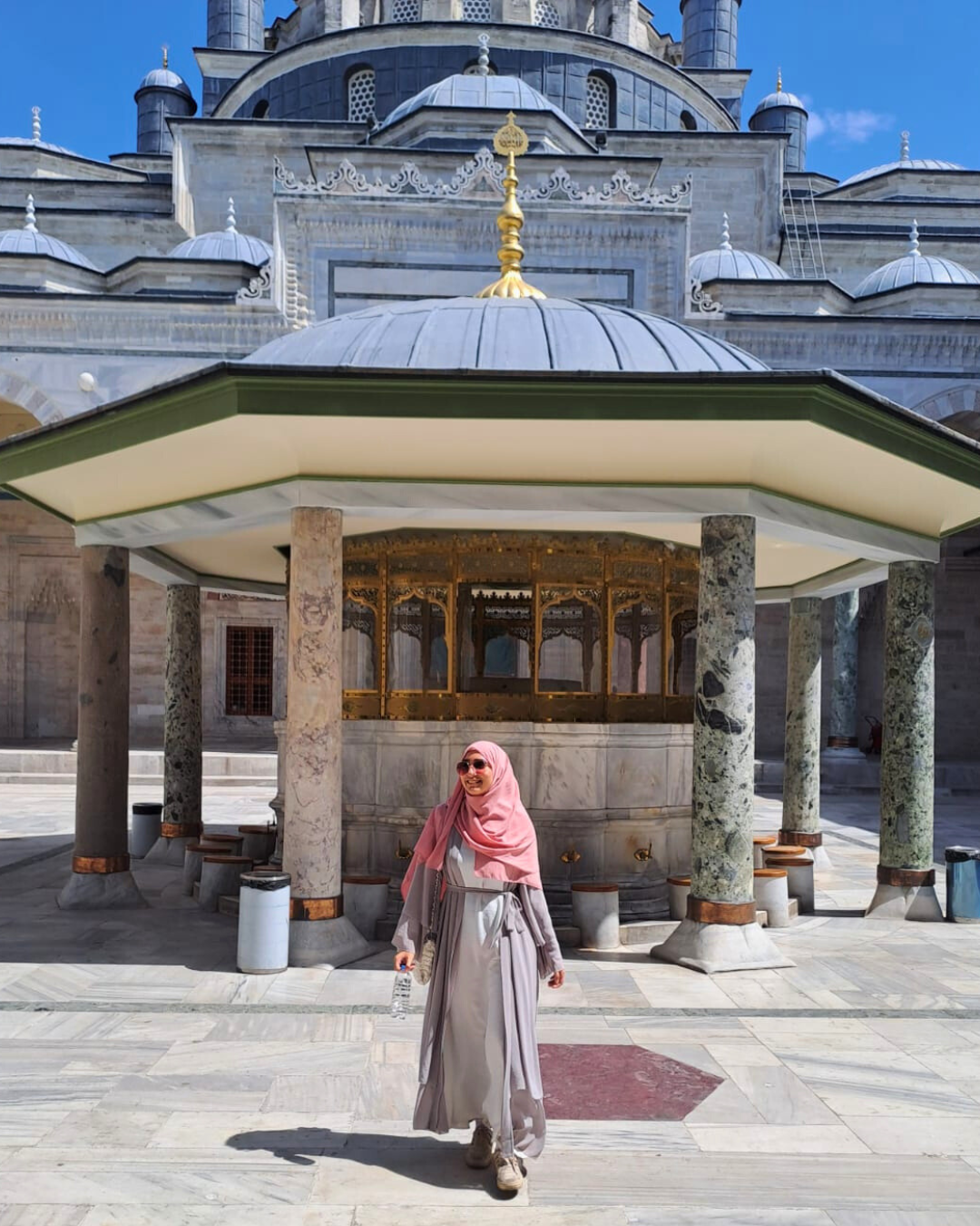 Sara is wearing a pink head scarf and sunglasses and is walking in front of a picturesque building in Istanbul, Turkey.