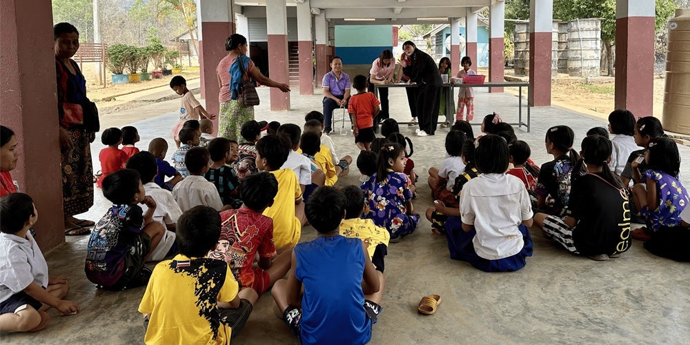 A group of children sit on the ground under a covered outdoor area facing a table where several adults are seated.