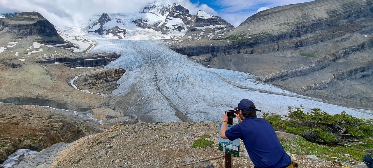 A man taking a photo at a lookout of a glacier with his phone.