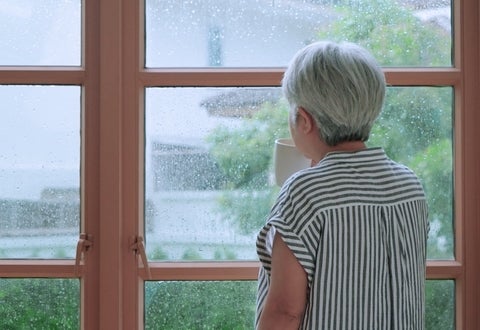 A senior person at home drinking from a cup while looking out a window.