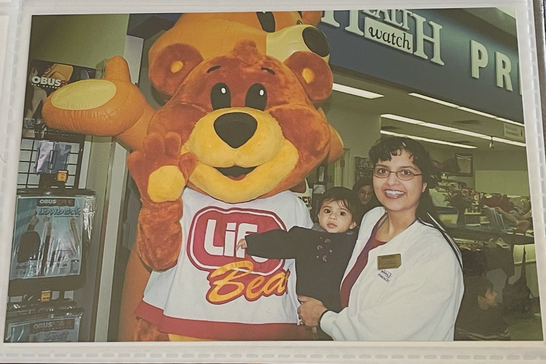 Baby Sapna Tripathi being held by her mom wearing pharmacist's lab coat with mascots