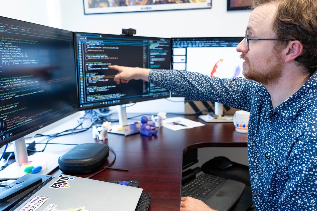 Joshua Pulsipher is seated and points at some code on the middle monitor of his three monitor set-up