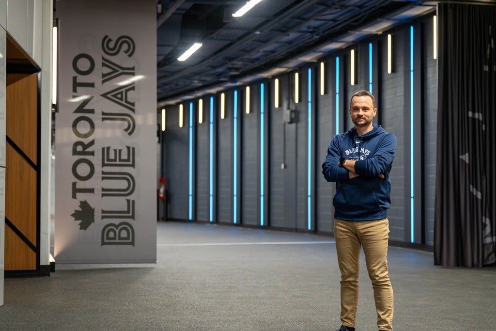 Jeremy Reesor stands with arms crossed outside blue jays clubhouse with signage and lighting behind him