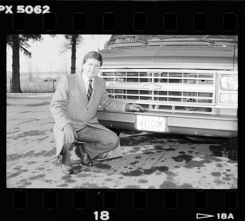 Ian McPhee kneeling beside a car with a license plate that reads "WATCOM"